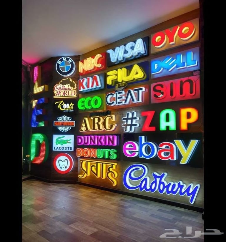 A large wall display of colorful illuminated neon brand logos including Visa, Nike, Fila, Dell, Kia, Eco, ARC, Dunkin Donuts, eBay, and Cadbury
